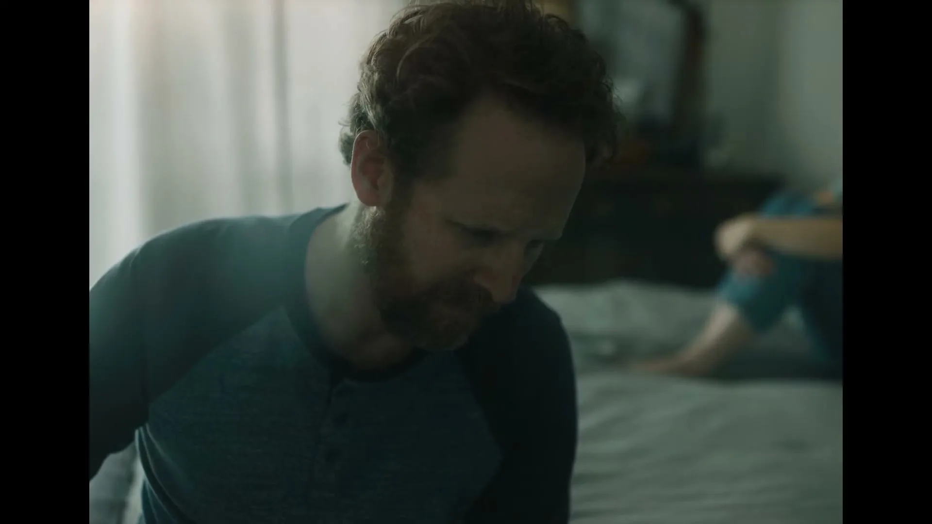 A man with a beard and blue shirt sits on a bed, looking down, while a woman sits in the background, in a dimly lit bedroom.