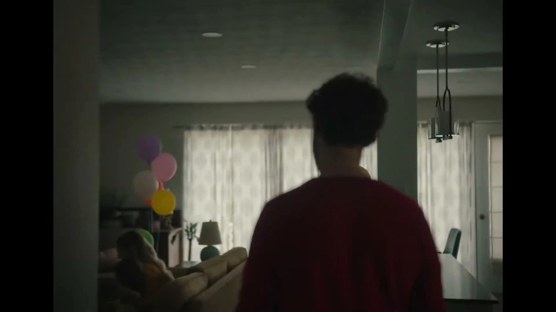 A man in a red shirt walks into a living room where a woman and child are sitting on a couch with balloons.