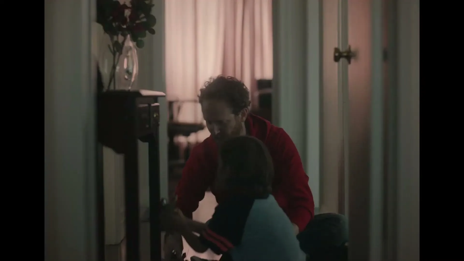 A father in a red shirt helps his young child put on a helmet in a dimly lit hallway, preparing for an activity.