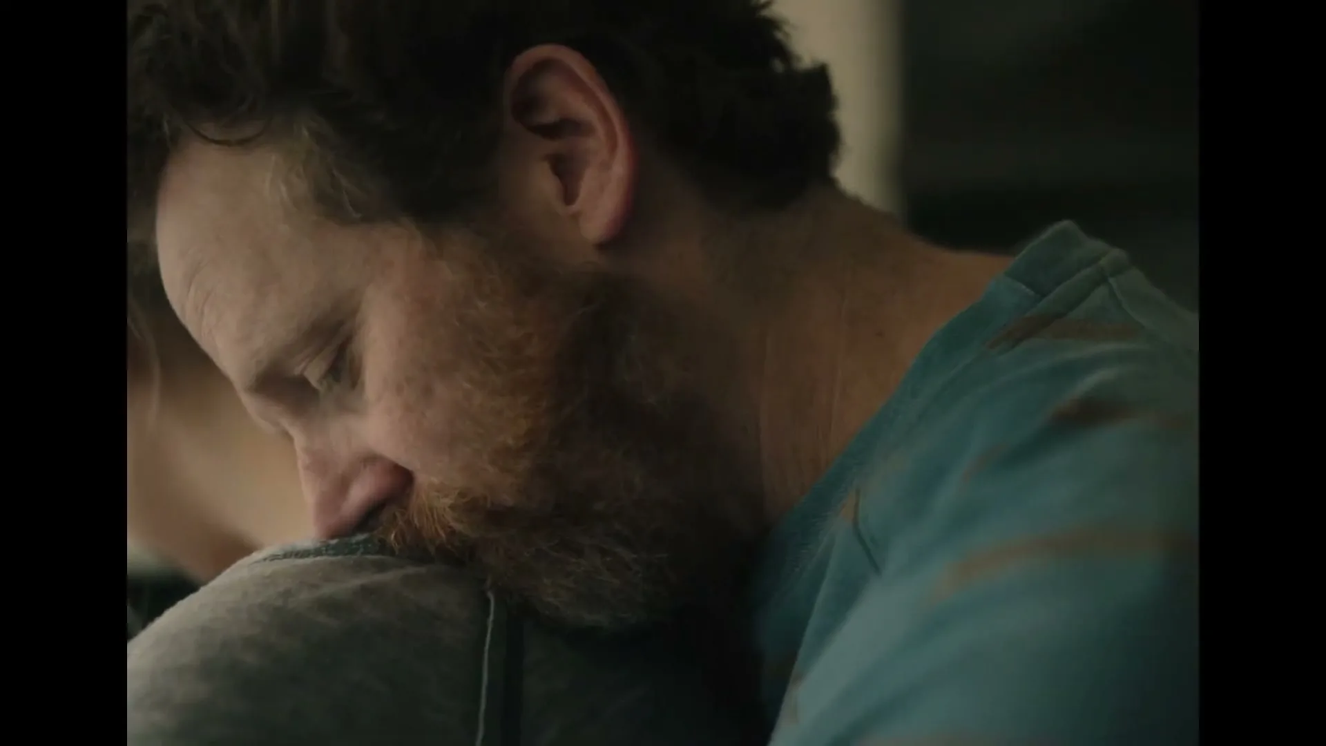 A man with a beard rests his head on a woman's shoulder, looking down with a somber expression in a dimly lit interior.