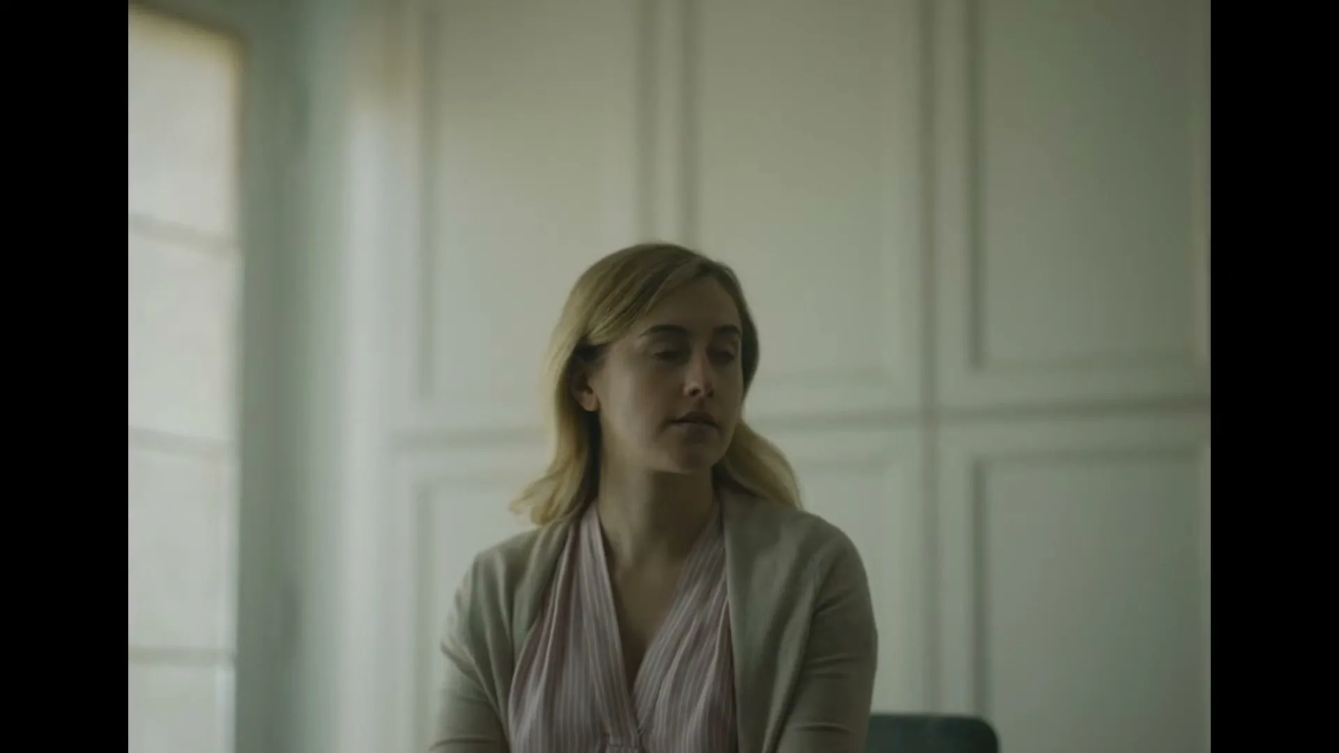 A man in a blue shirt turns away from the camera, then a woman with blonde hair in a pink shirt and beige cardigan looks down, in a bright room.
