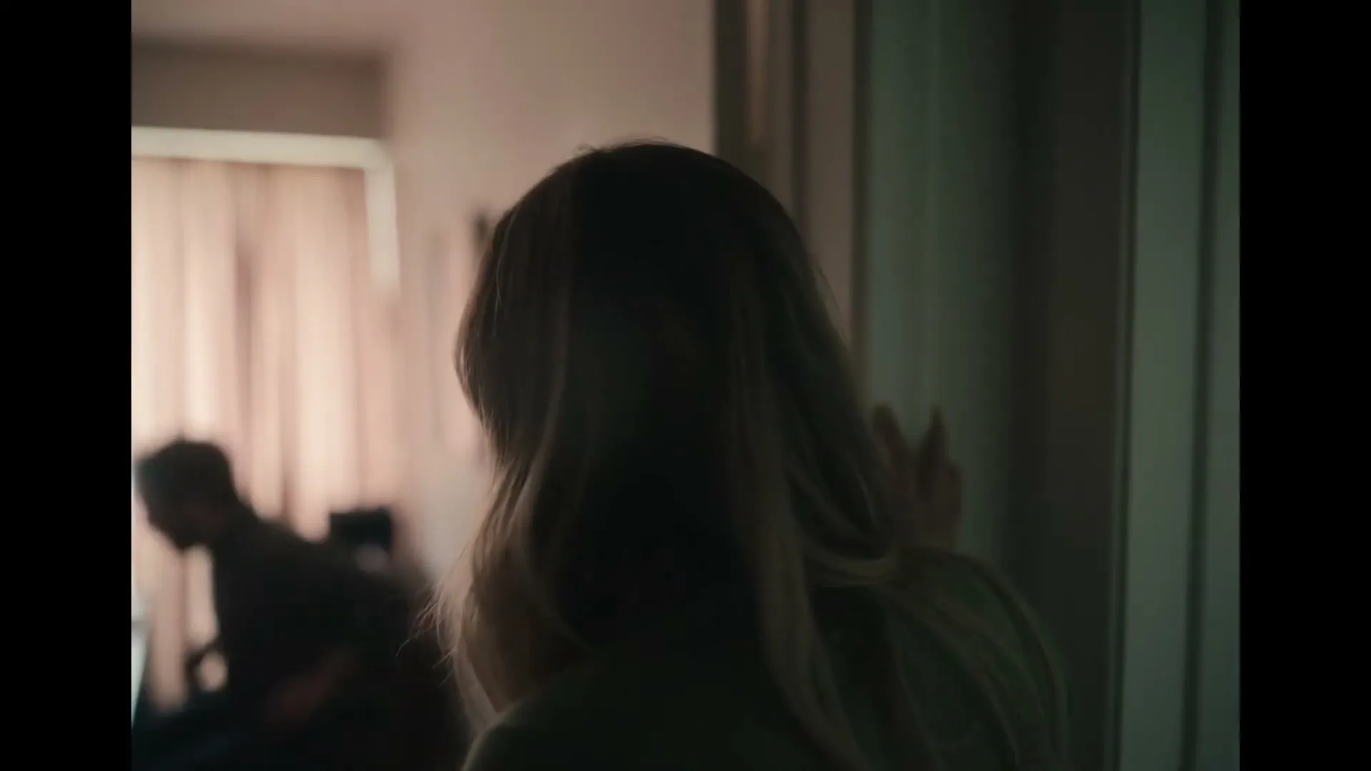 A woman with long blonde hair, seen from behind, stands in a doorway as a man walks past in the background, in a dimly lit room.