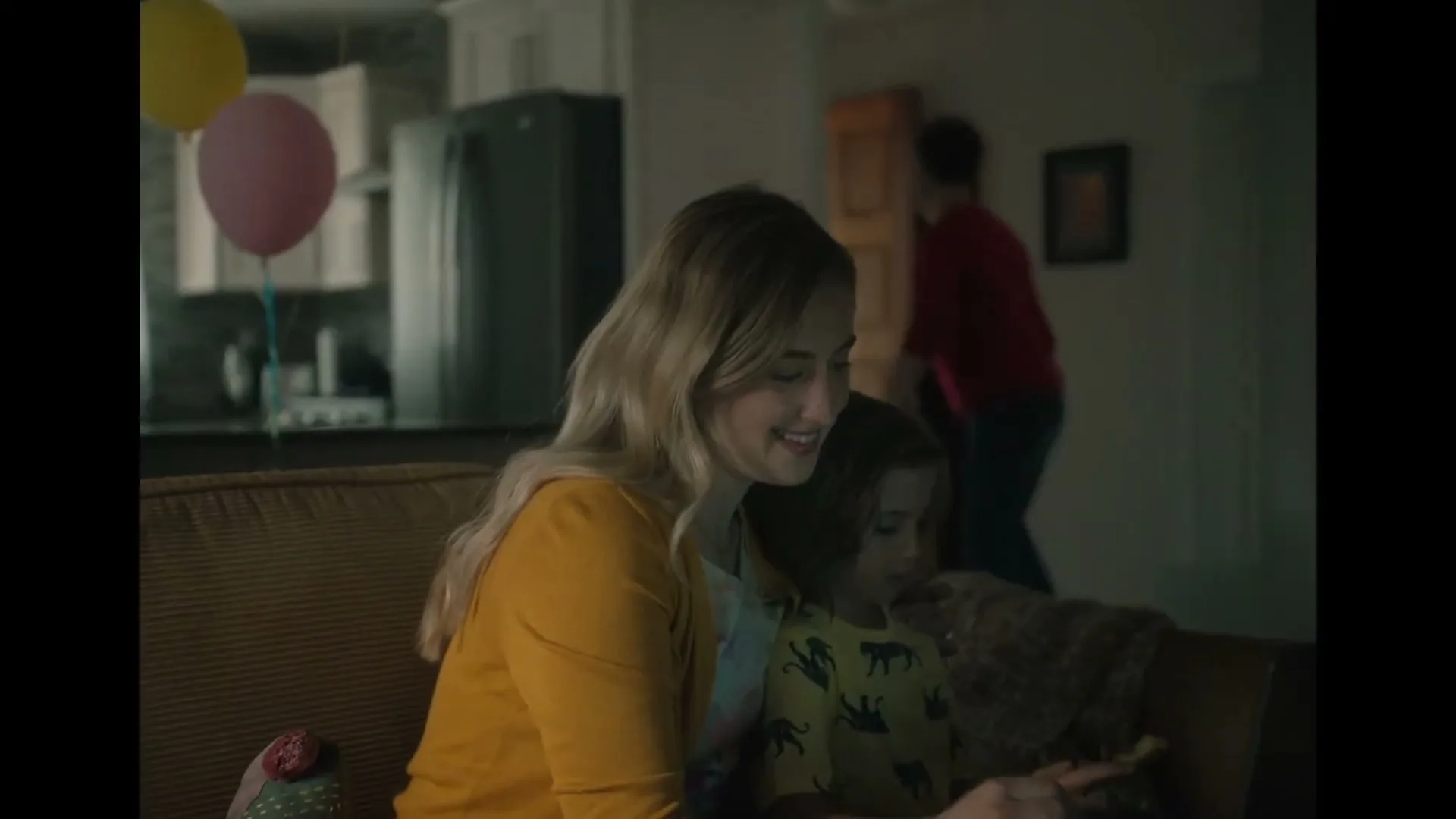 A smiling woman in a yellow cardigan sits on a couch with a child, looking at a phone, while a man walks in the background of a living room.
