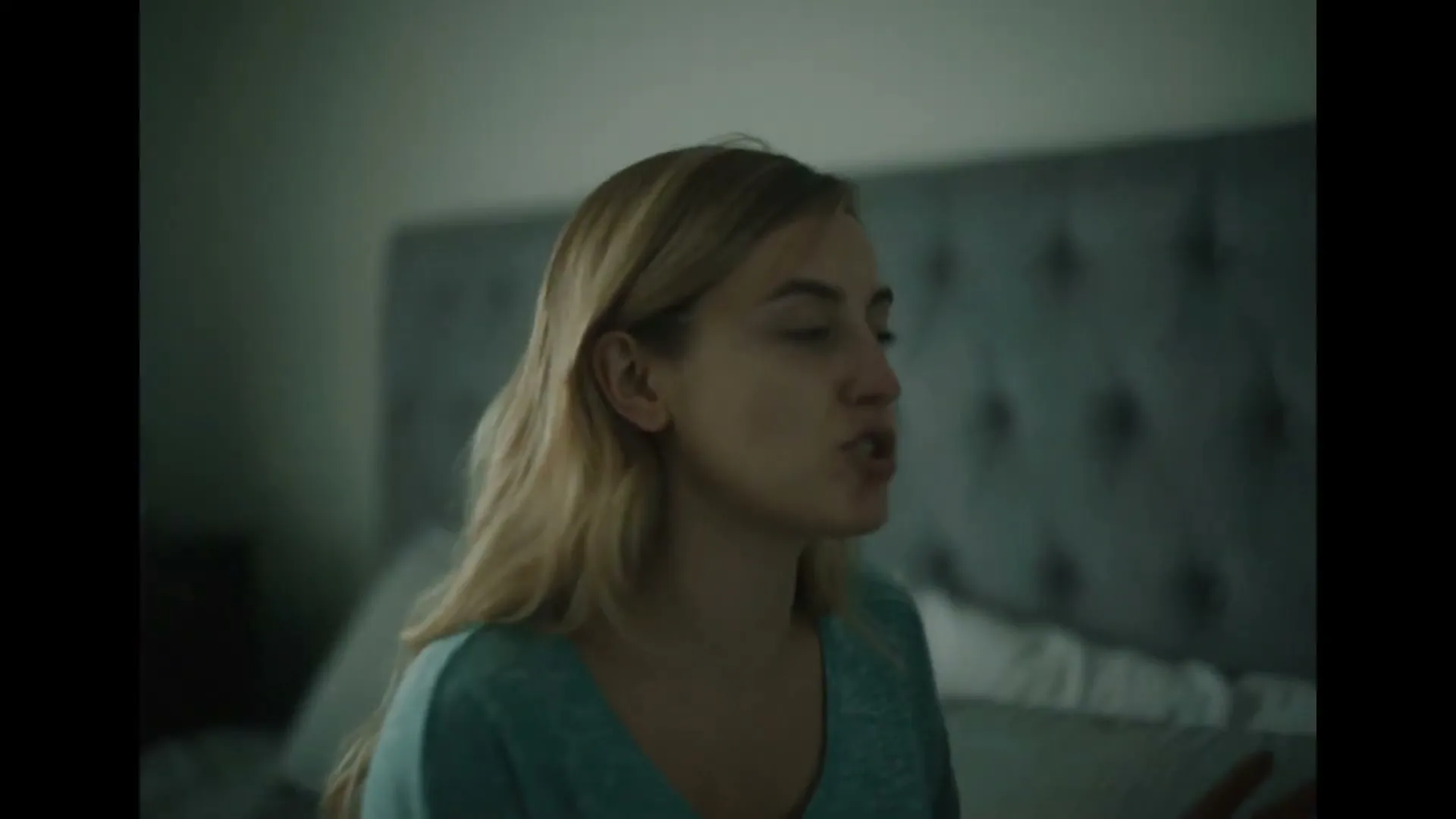 A young woman with blonde hair in a teal shirt argues with an unseen person, gesturing with her hands in a dimly lit bedroom.