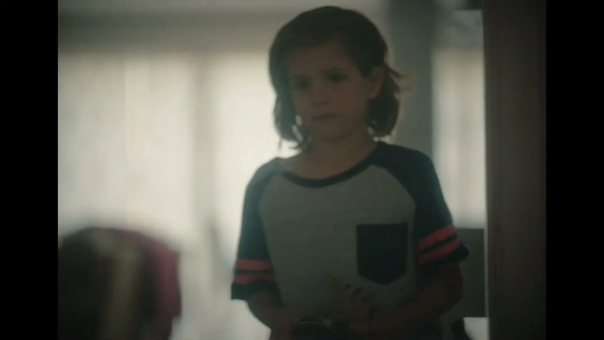 A young child with shoulder-length hair, wearing a two-toned t-shirt, looks down sadly in a softly lit room.