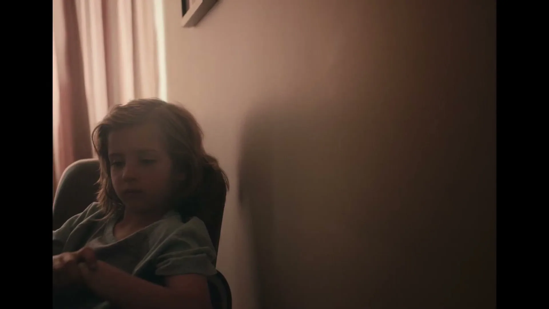 A young child with long hair sits in a chair, looking down with a somber expression, in a dimly lit room with a plain wall and curtains.