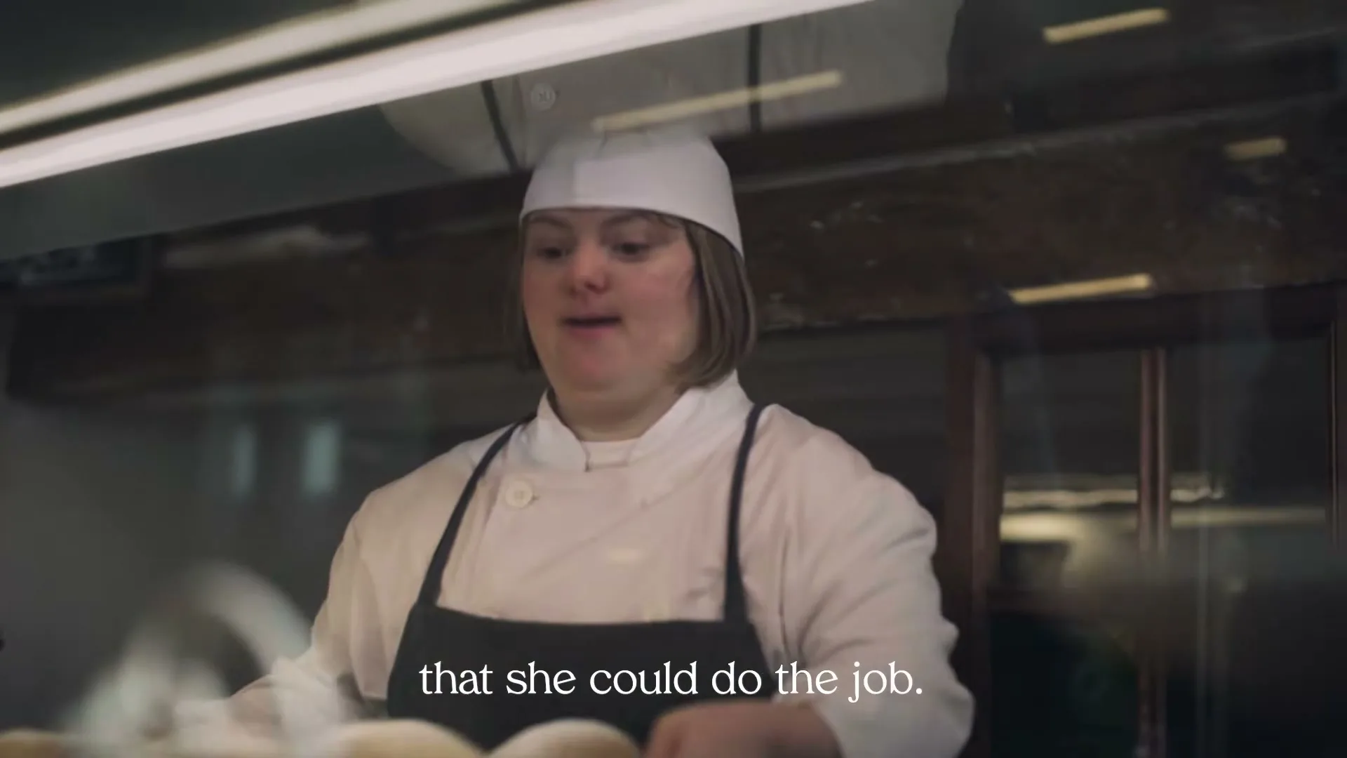 A young woman with Down syndrome, wearing a chef's hat and apron, places a tray of baked goods into a display case in a bakery.