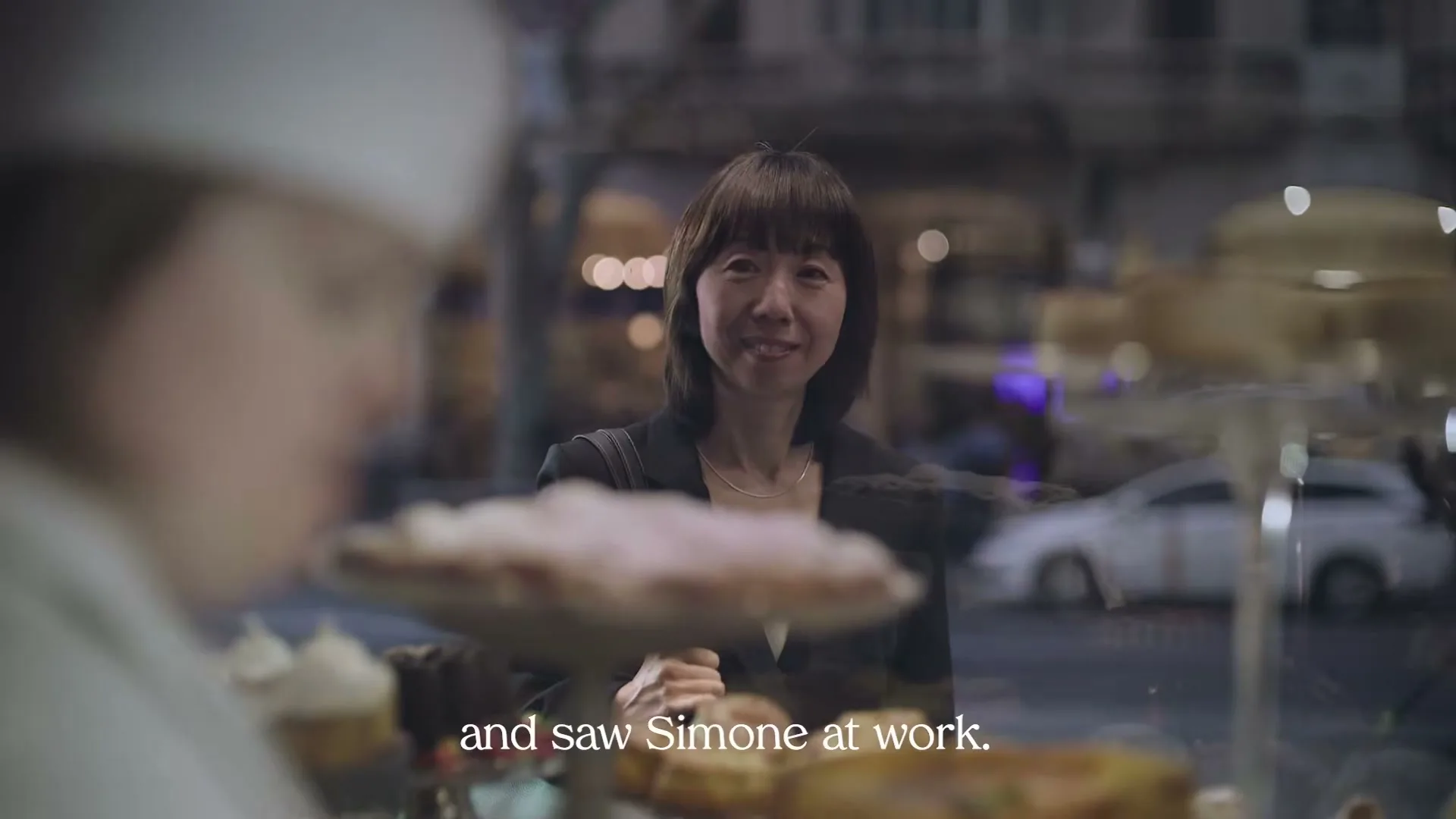 A woman with dark hair and a black blazer smiles while looking through a bakery window at a baker in a white hat.