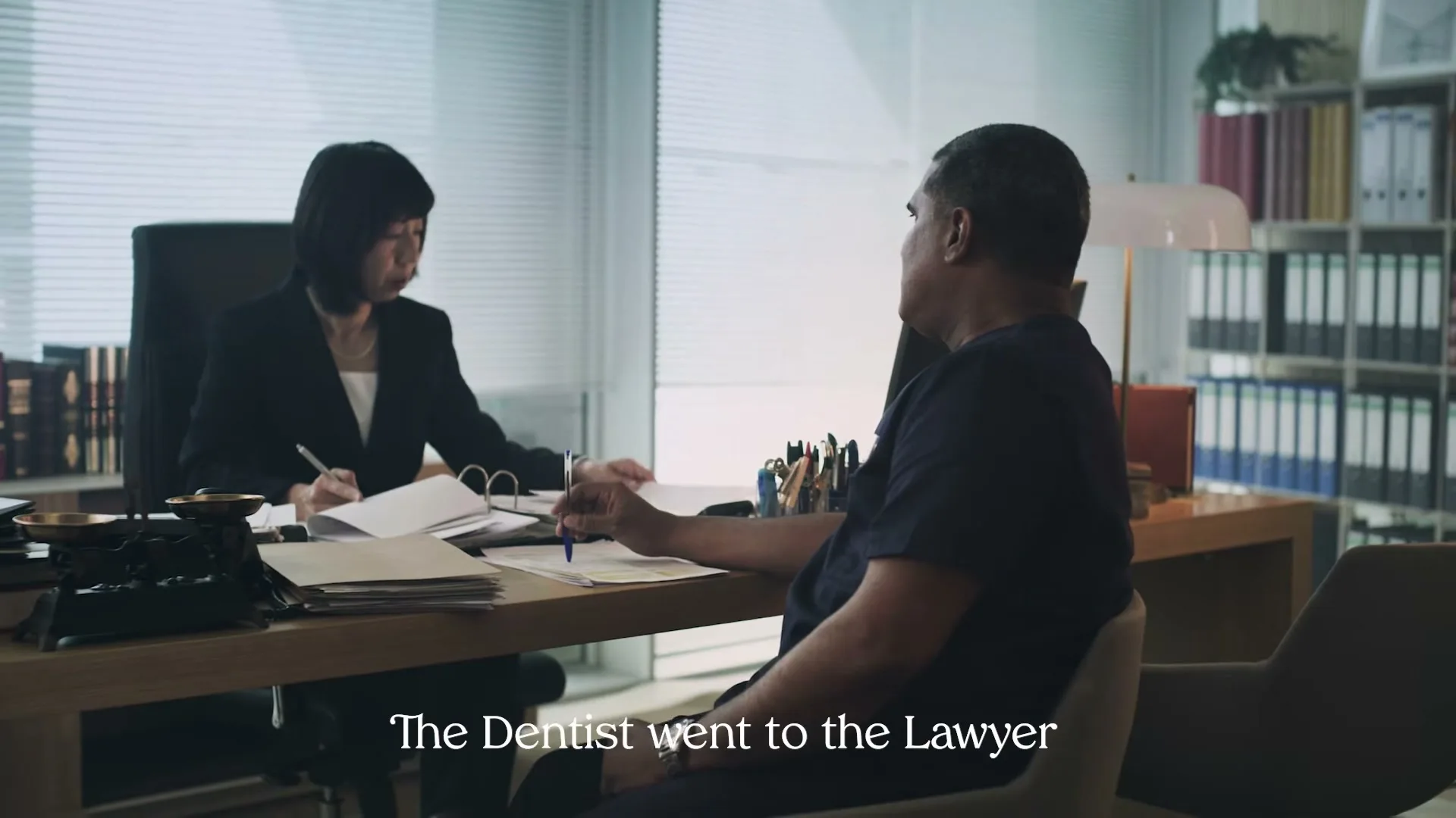 A male dentist in a dark blue scrub top sits at a desk, looking at a female lawyer in a black suit who is writing on papers in an office.