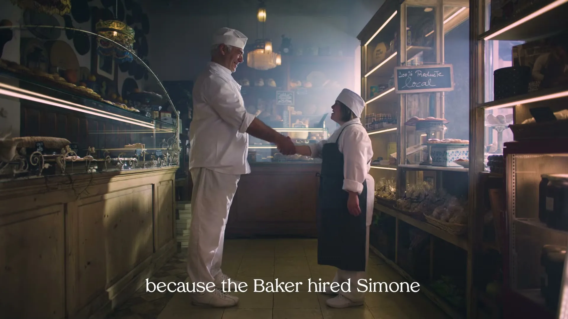A male baker in his 60s and a female baker with Down syndrome in her 20s shake hands in a warmly lit bakery.