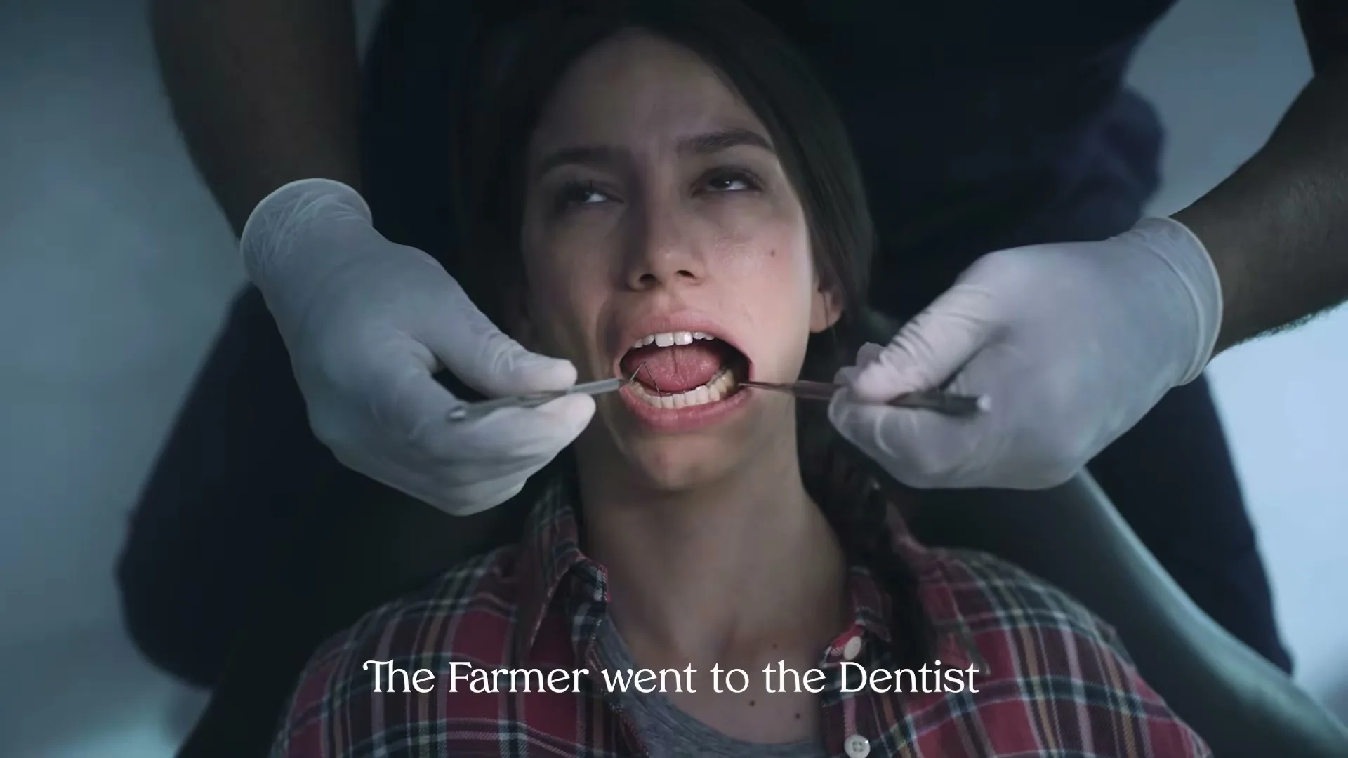 A young woman with dark hair, wearing a plaid shirt, is seen in a dentist's chair with her mouth open, as a dentist's gloved hands hold tools inside her mouth.