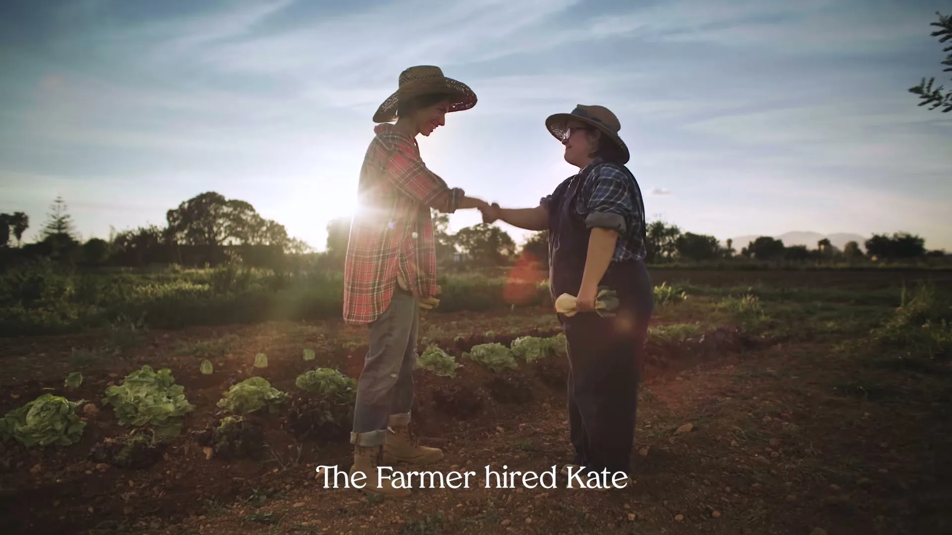 A female farmer in a plaid shirt and straw hat shakes hands with a woman with Down syndrome in overalls and a straw hat, in a sunny field.
