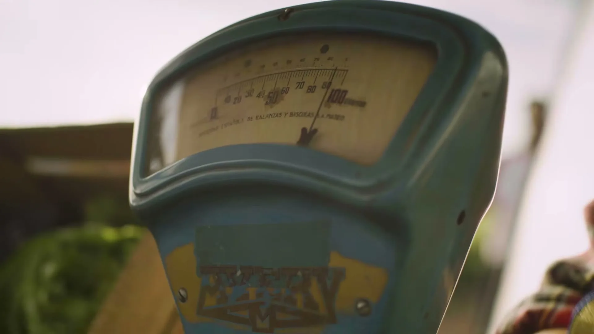 A close-up shot of an old, teal-colored weighing scale with a dial showing numbers, at an outdoor market.