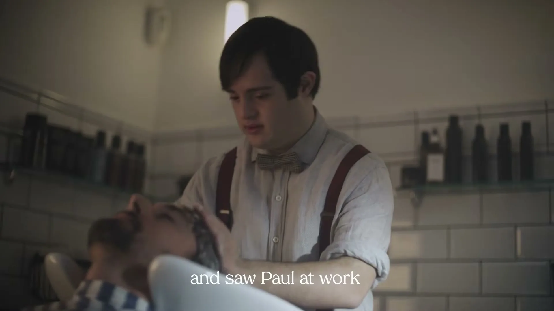 A young man with Down syndrome, wearing a shirt and suspenders, washes the hair of a male customer in a barbershop.