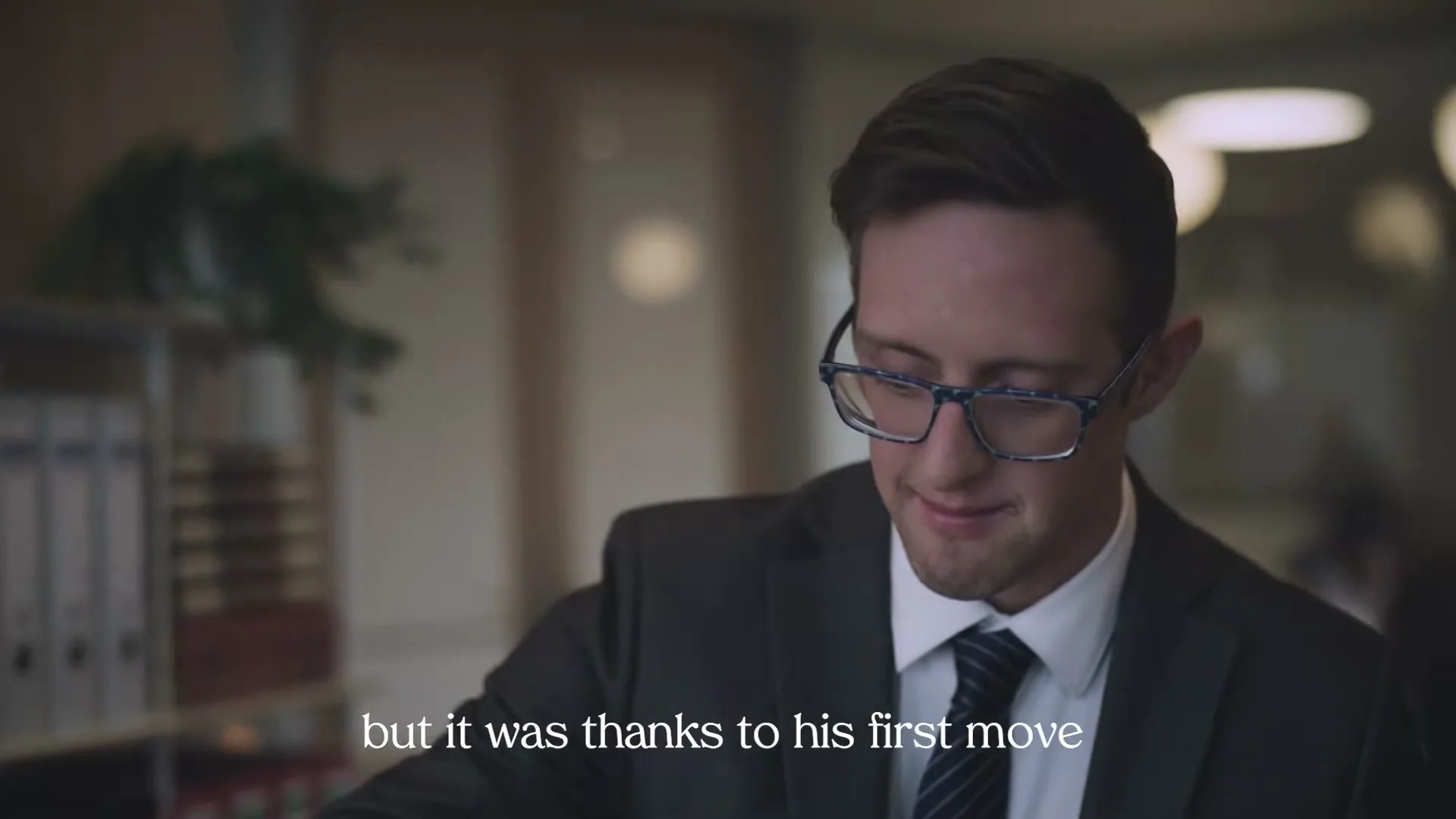 A young man with Down syndrome, wearing a suit and glasses, looks down and smiles shyly in an office setting.