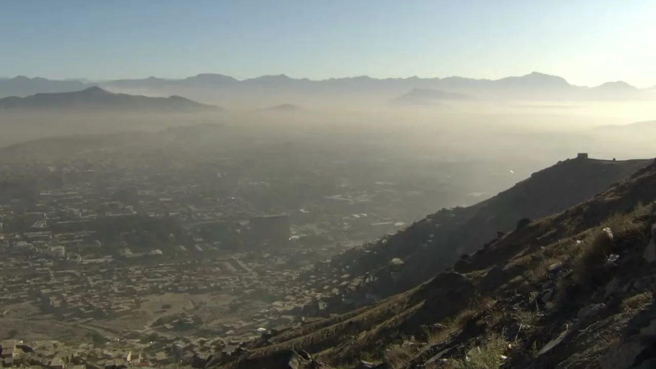 An extreme wide shot shows a hazy, sprawling city nestled in a valley, surrounded by rugged mountains under a clear sky.