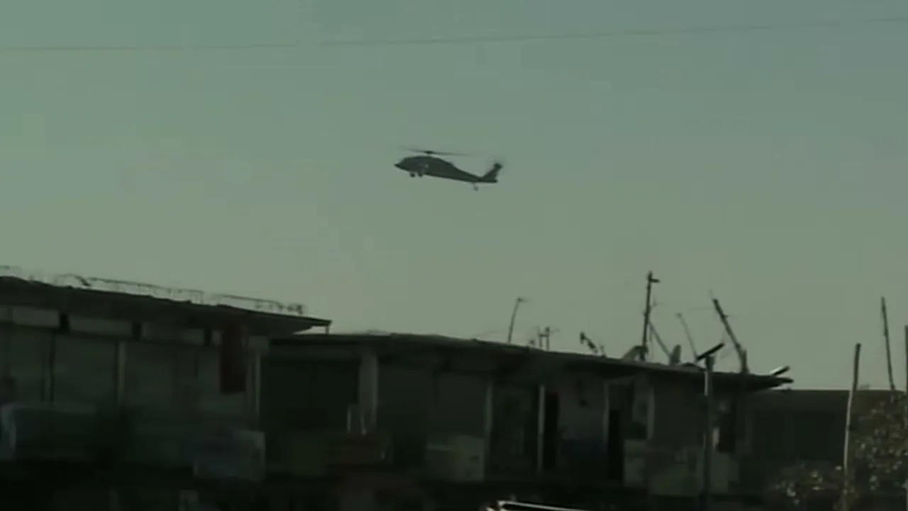 A military helicopter flies across a hazy sky above dilapidated buildings in a war-torn area during the day.