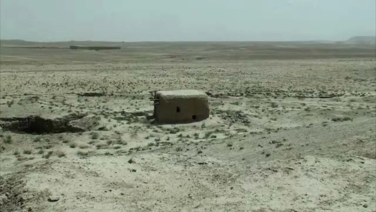 A small, dilapidated mud hut stands alone in a vast, arid desert landscape, then explodes into a cloud of dust and debris.