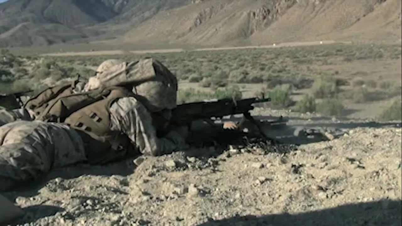 A soldier in camouflage uniform lies prone on a rocky desert ground, firing a machine gun with mountains in the background.