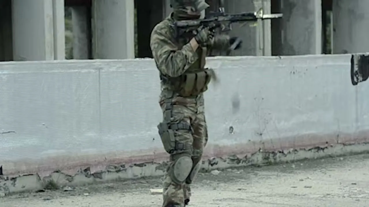 A soldier in camouflage uniform and tactical gear reloads a rifle in a desolate, war-torn urban environment.