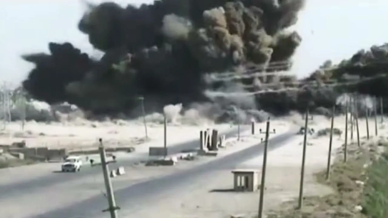 A massive explosion erupts on the side of a desert road, sending a huge cloud of black smoke and debris into the air, with a white car driving away.