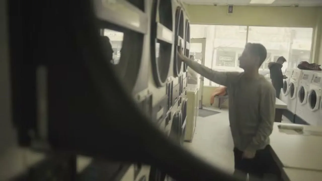A young man in a long-sleeved shirt and dark pants is seen from a medium-wide shot, standing in a laundromat and closing a washing machine door.