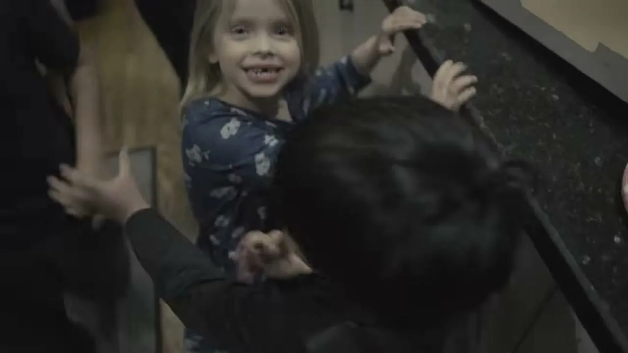 A young girl with blonde hair and a missing front tooth smiles at the camera while holding onto a kitchen counter, with another child's head in the foreground.
