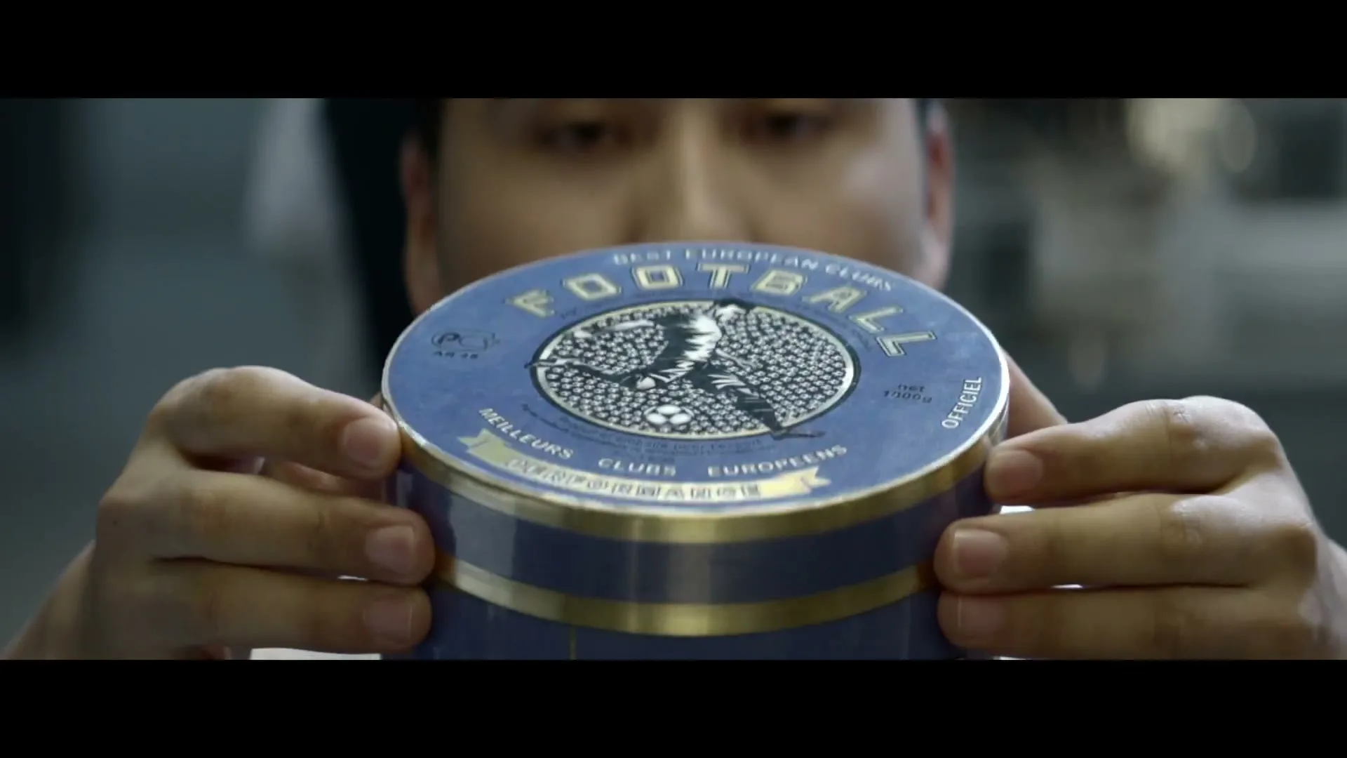 A man in a white chef's coat holds a blue and gold tin of "Football" branded caviar, looking at it intently in a kitchen.