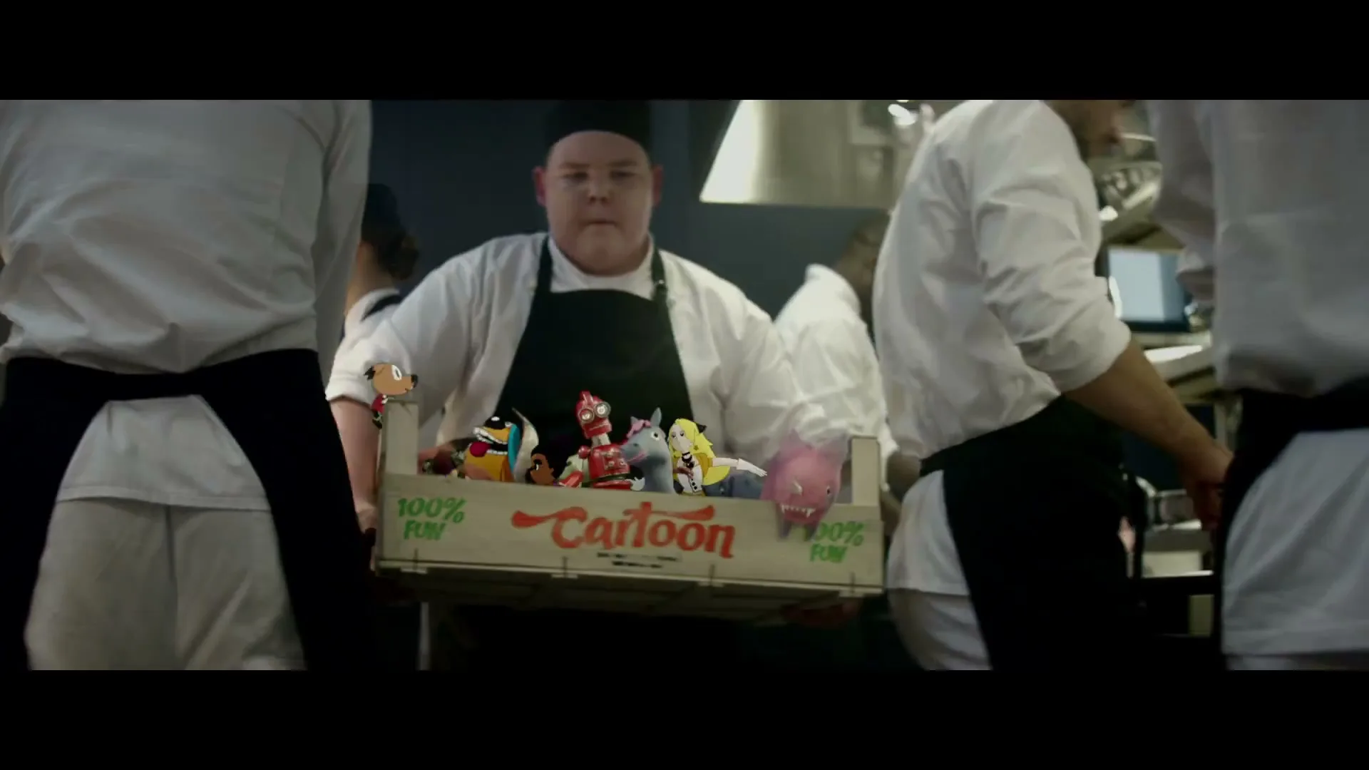 A male chef in a white uniform and black apron carries a wooden crate filled with animated cartoon characters through a busy kitchen.