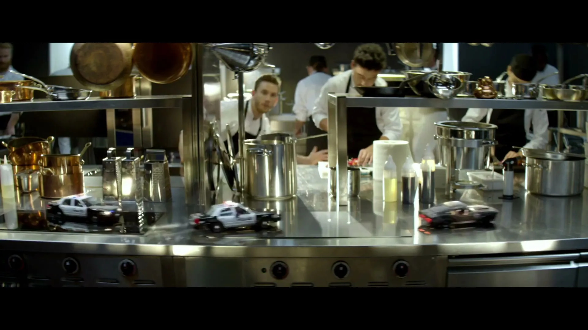Miniature police cars drive across a busy, stainless steel kitchen counter where chefs are working in the background.