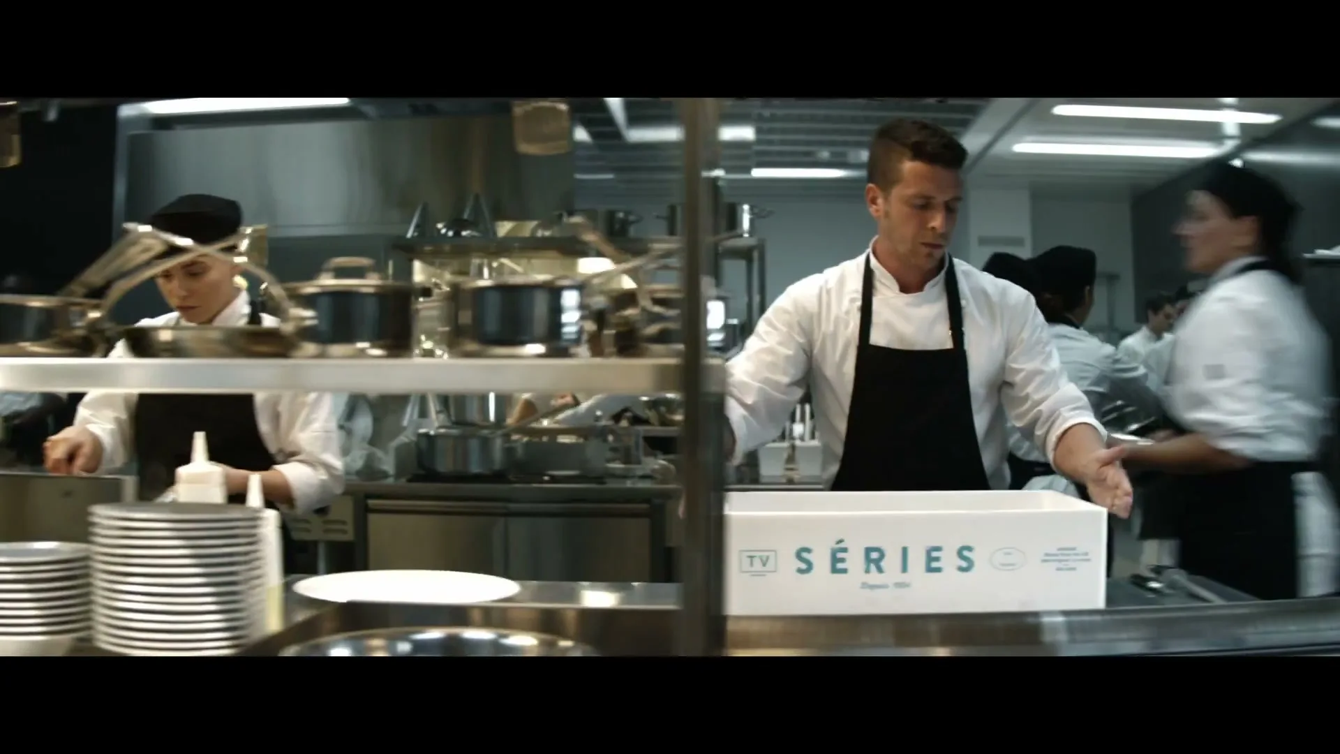 A male chef in a white uniform and black apron places a white box with "TV SERIES" on it onto a metal counter in a busy commercial kitchen.