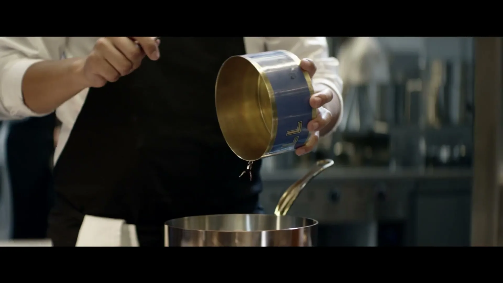 A chef in a black apron and white shirt holds a blue and gold tin, pouring liquid into a pot in a professional kitchen.