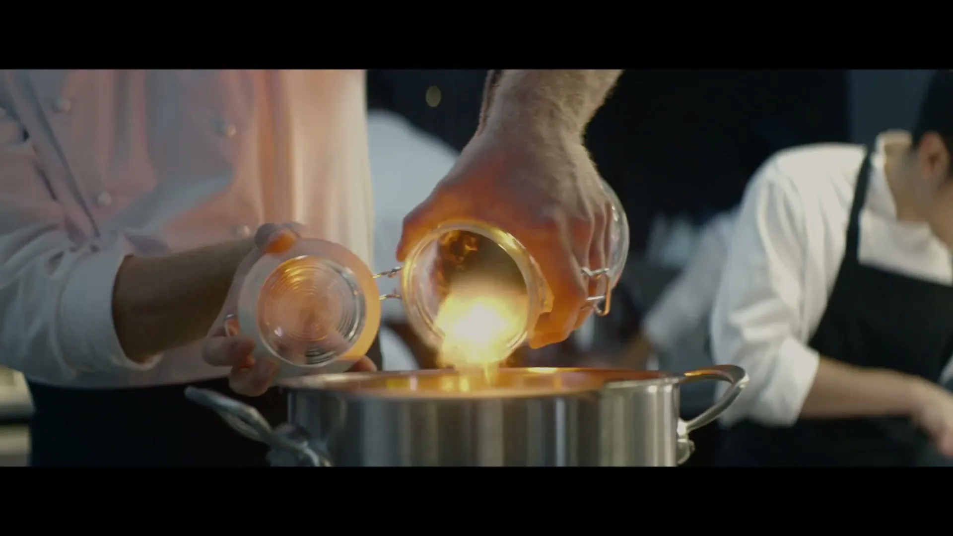 A chef's hands pour glowing ingredients from a glass jar into a pot, with another chef in the background.