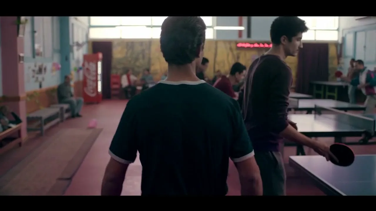 A young man in a dark t-shirt walks through a busy community hall with people playing table tennis and sitting around.