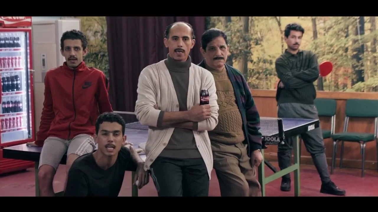 Five men of varying ages are gathered around a ping-pong table in a community hall, looking directly at the camera with serious expressions.