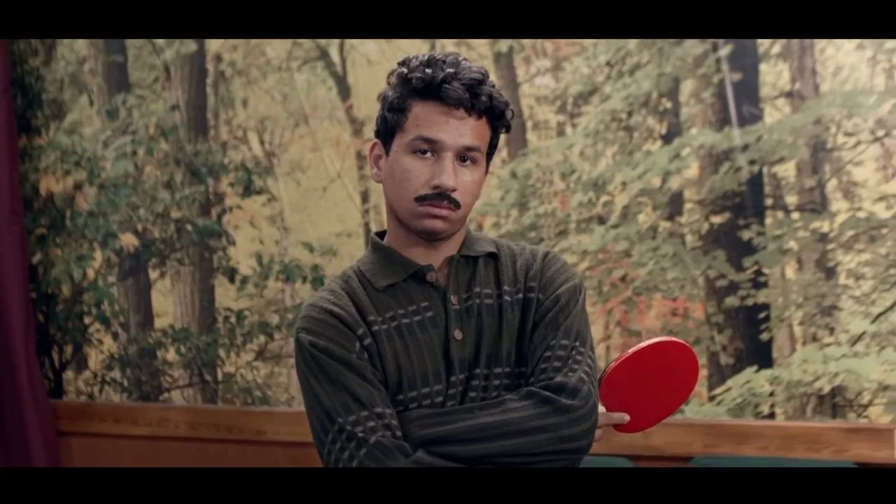 A young man with a mustache, wearing a green sweater, stands with arms crossed, holding a red ping-pong paddle, in a room with a forest mural.
