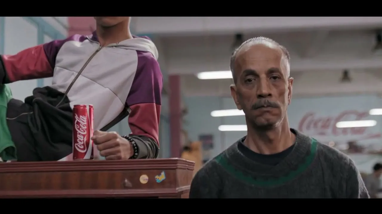 A man with a mustache looks directly at the camera, while a younger man with a drawn-on mustache sits behind him with a Coca-Cola can.