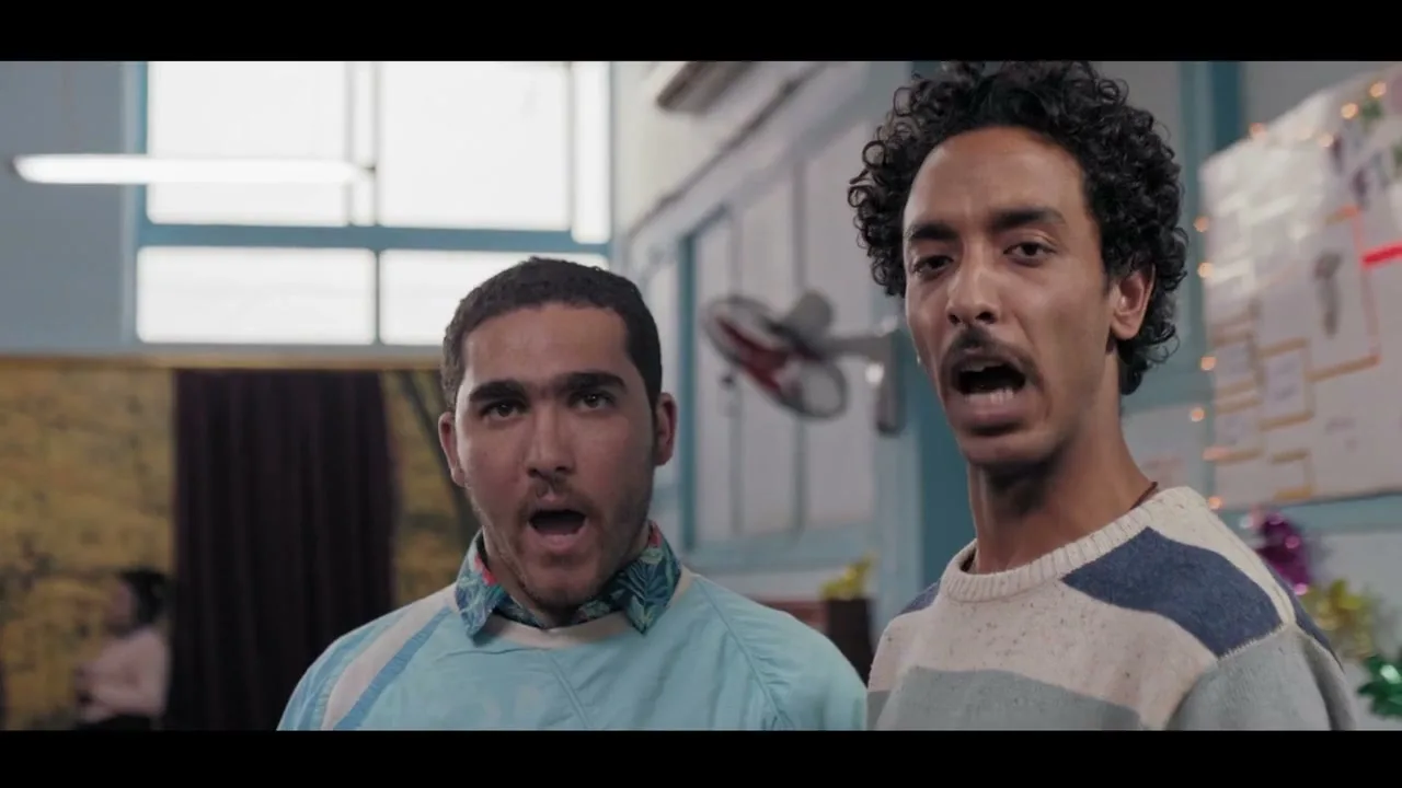 Two young men, one with a mustache and curly hair, the other with a beard, stand side-by-side, looking forward with open mouths in an interior setting.