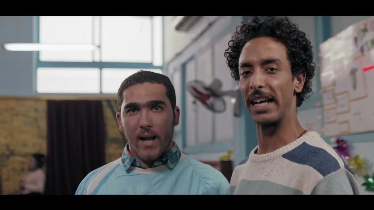 Two young men, one with a mustache and curly hair, the other with a beard, stand side-by-side, looking forward with open mouths in an interior setting.