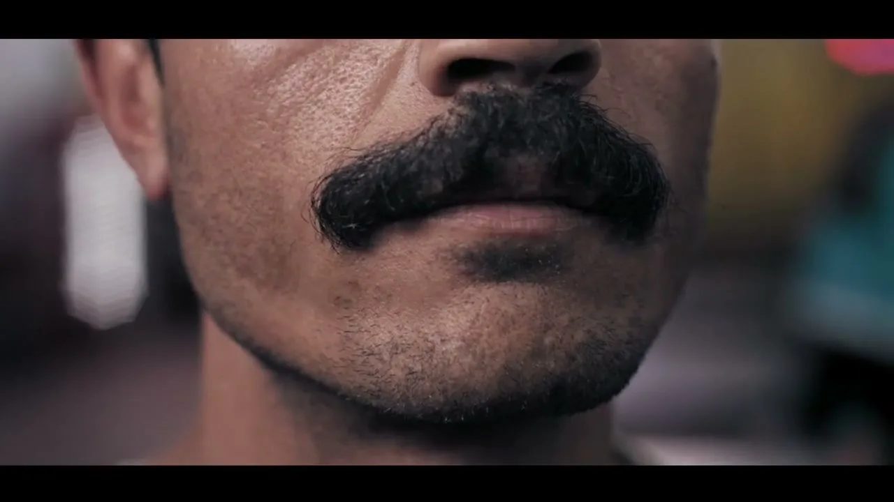 A man with a thick black mustache and stubble on his chin looks forward, his mouth slightly open, in a medium closeup shot.