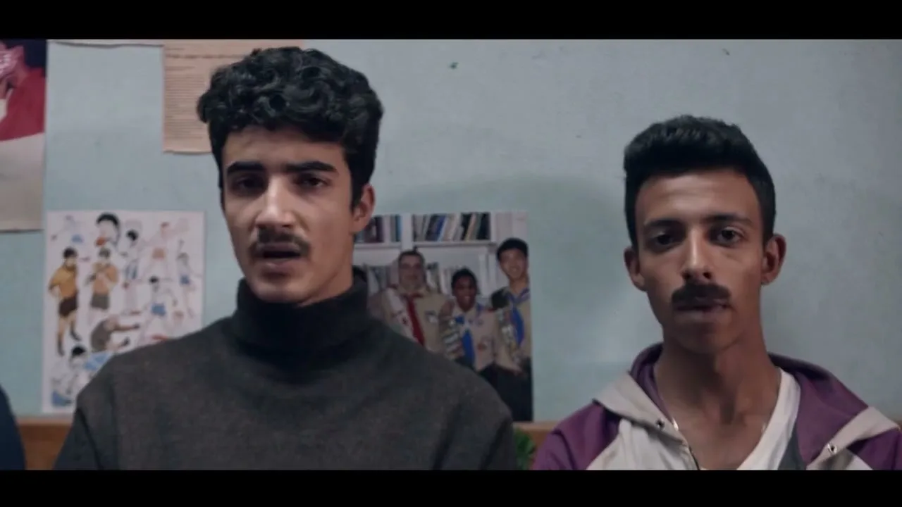 Two young men with mustaches look surprised and then directly at the camera in an interior setting.