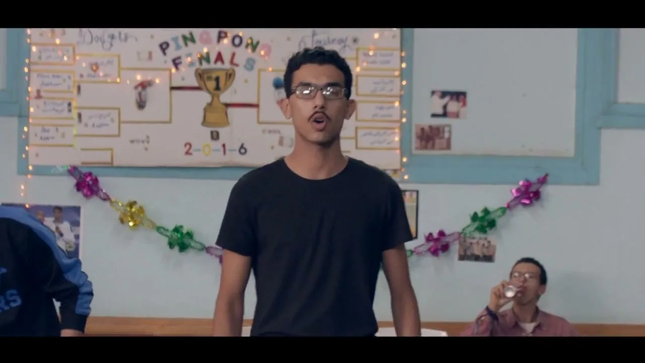 A young man with glasses and a mustache sings into the camera, while two other men play ping pong and another watches in a community hall.