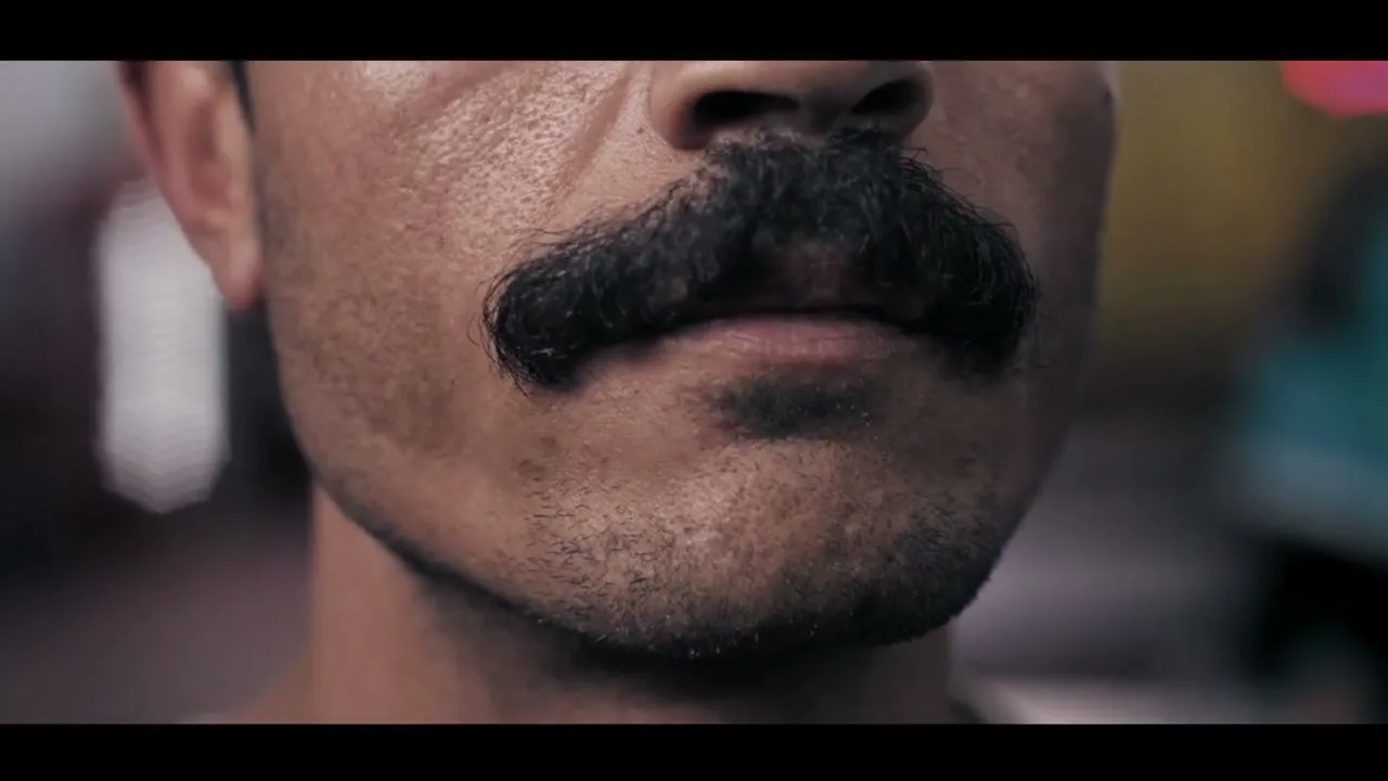A man with a thick black mustache and stubble on his chin is shown in a close-up shot, looking directly at the camera with a neutral expression.