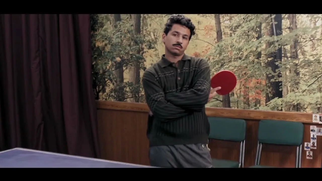 A man with a mustache, wearing a green sweater and grey shorts, stands with arms crossed, holding a red ping-pong paddle in a room with a forest mural.