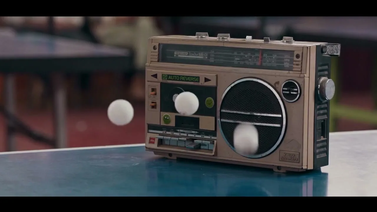 A vintage boombox sits on a blue ping-pong table as white ping-pong balls bounce around it in an interior setting.