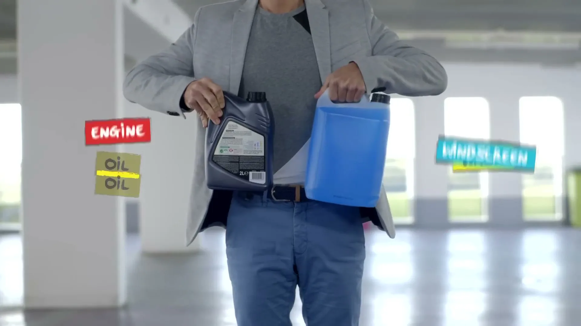 A man in a grey blazer and blue pants stands in a bright, open space, revealing engine oil and windscreen wash from behind his back.