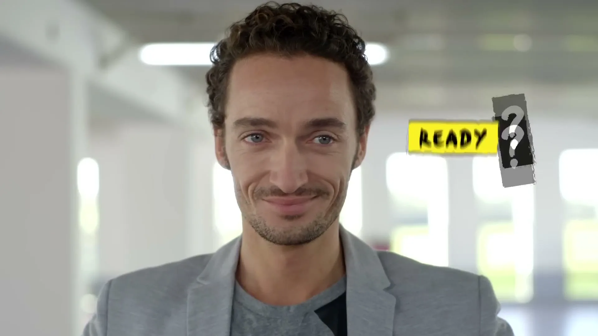 A man with curly brown hair and a light grey blazer smiles subtly at the camera, with "READY" and a question mark appearing next to him.