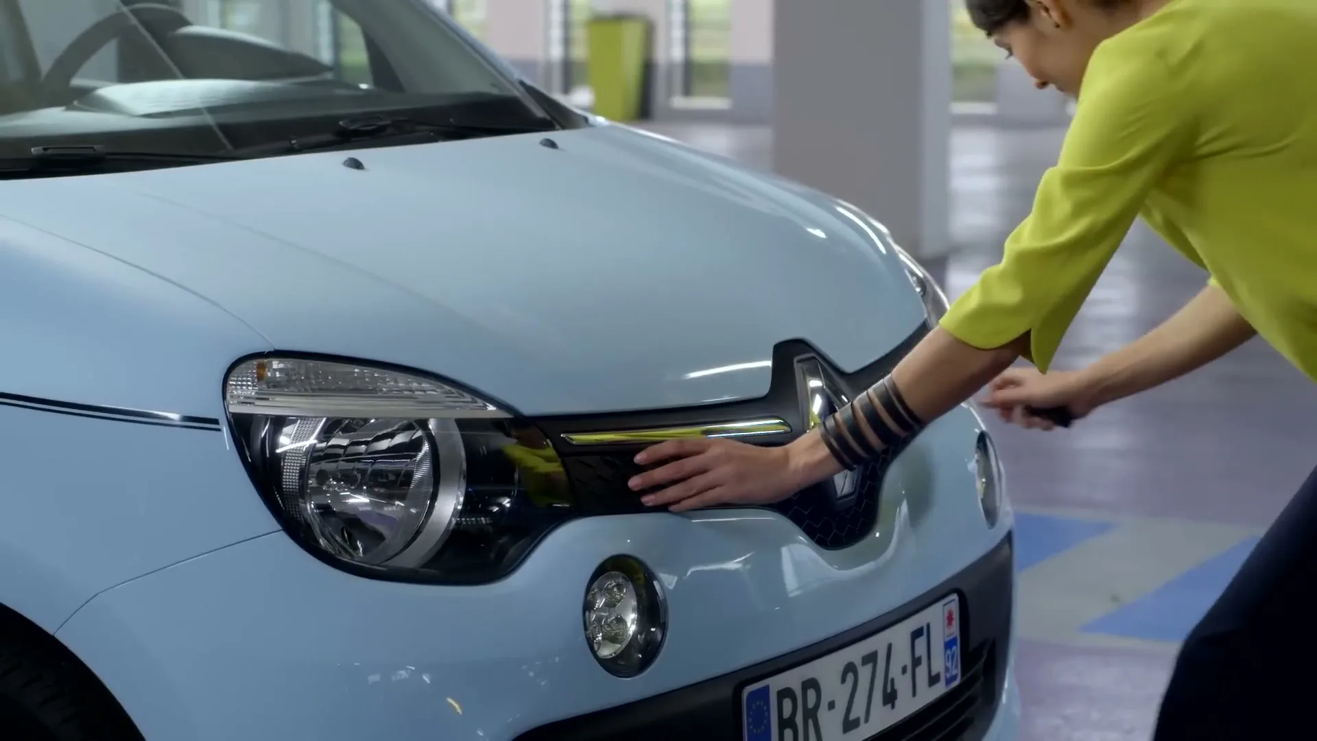 A woman in a yellow shirt and dark pants opens the hood of a light blue Renault Twingo in an underground parking garage.