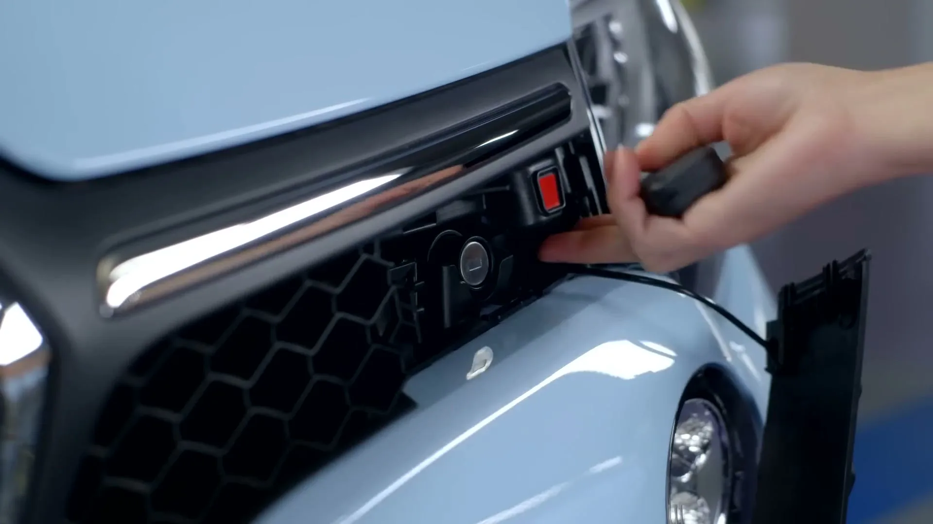 A person's hand inserts a key into a light blue car's front grille, revealing a hidden compartment.