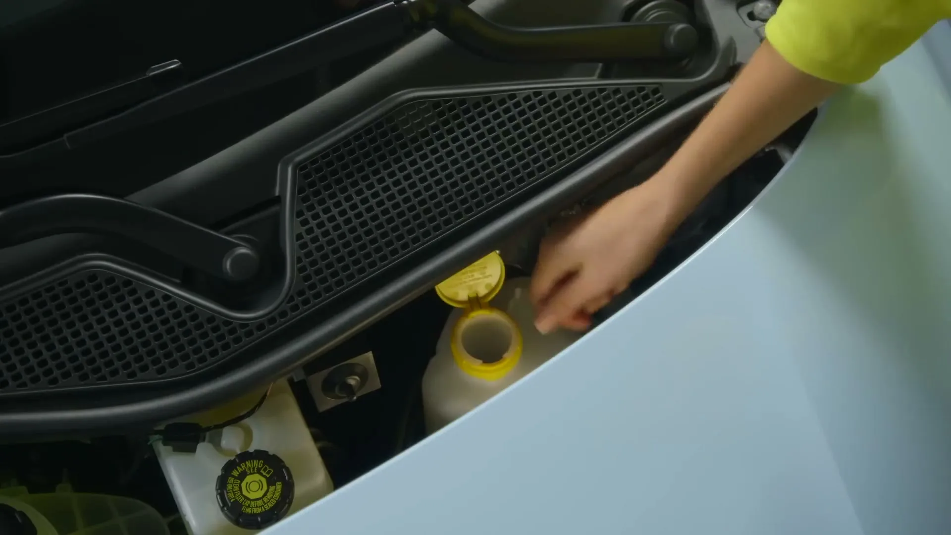 A person's hand in a yellow shirt opens and closes the cap of a fluid reservoir in the engine bay of a light blue car.